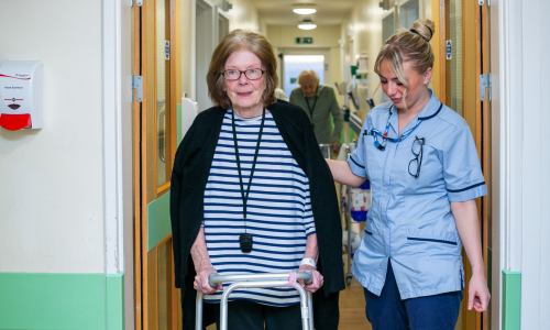 Member of staff helping patient with walking equipment walk down the hospital corridor